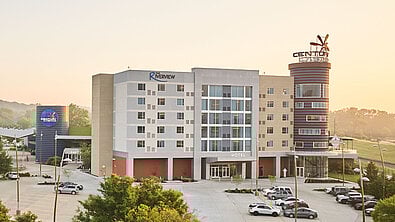 A modern hotel building with multiple floors, featuring a colorful sign and a spacious parking lot. Trees and hills are in the background.